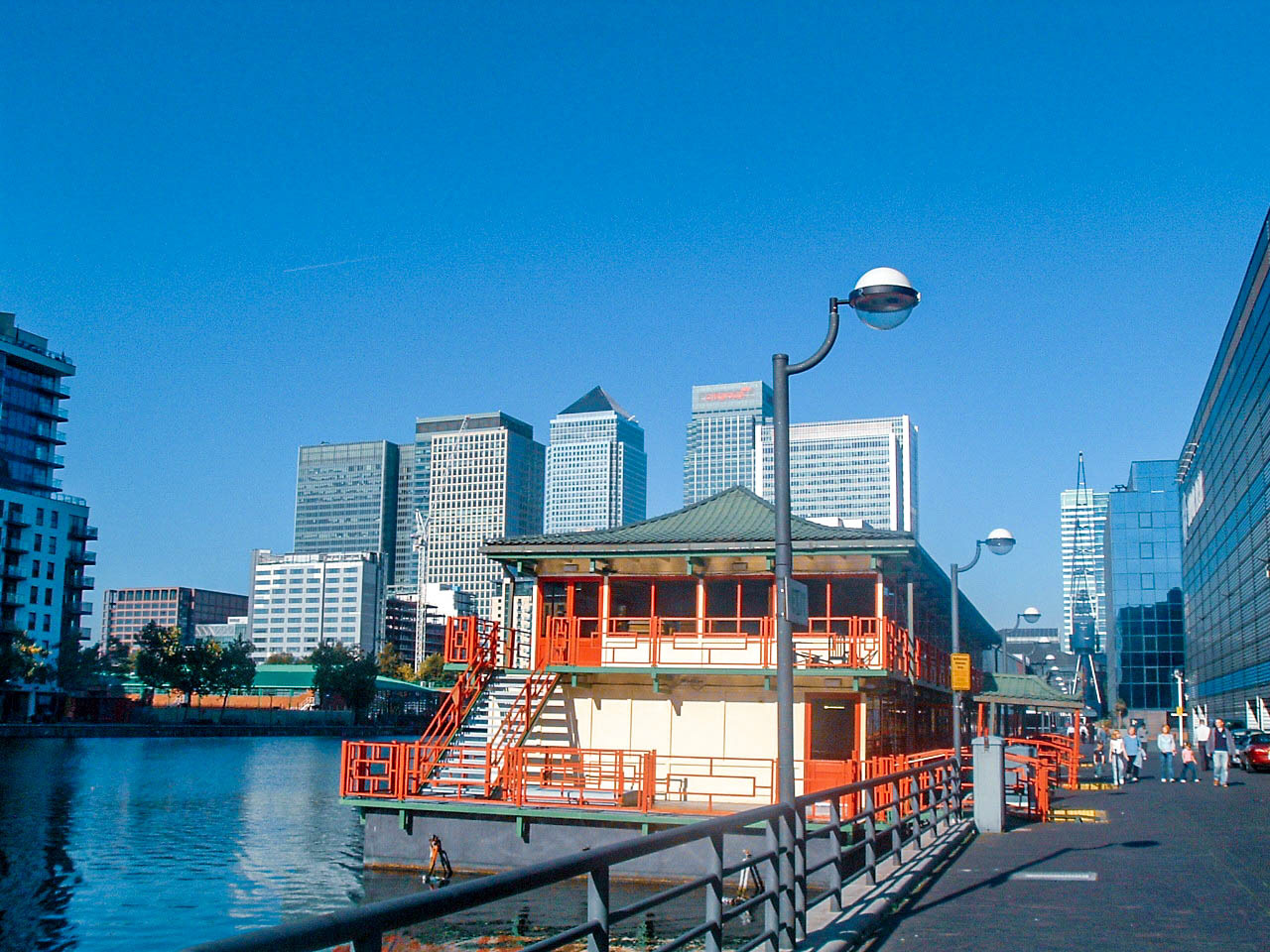 The image depicts a modern urban waterfront scene with a traditional-style building in the foreground, surrounded by contemporary high-rise buildings. The scene includes a calm body of water, a pedestrian walkway with railings, and several people walking along the path. The sky is clear and blue, suggesting a sunny day.