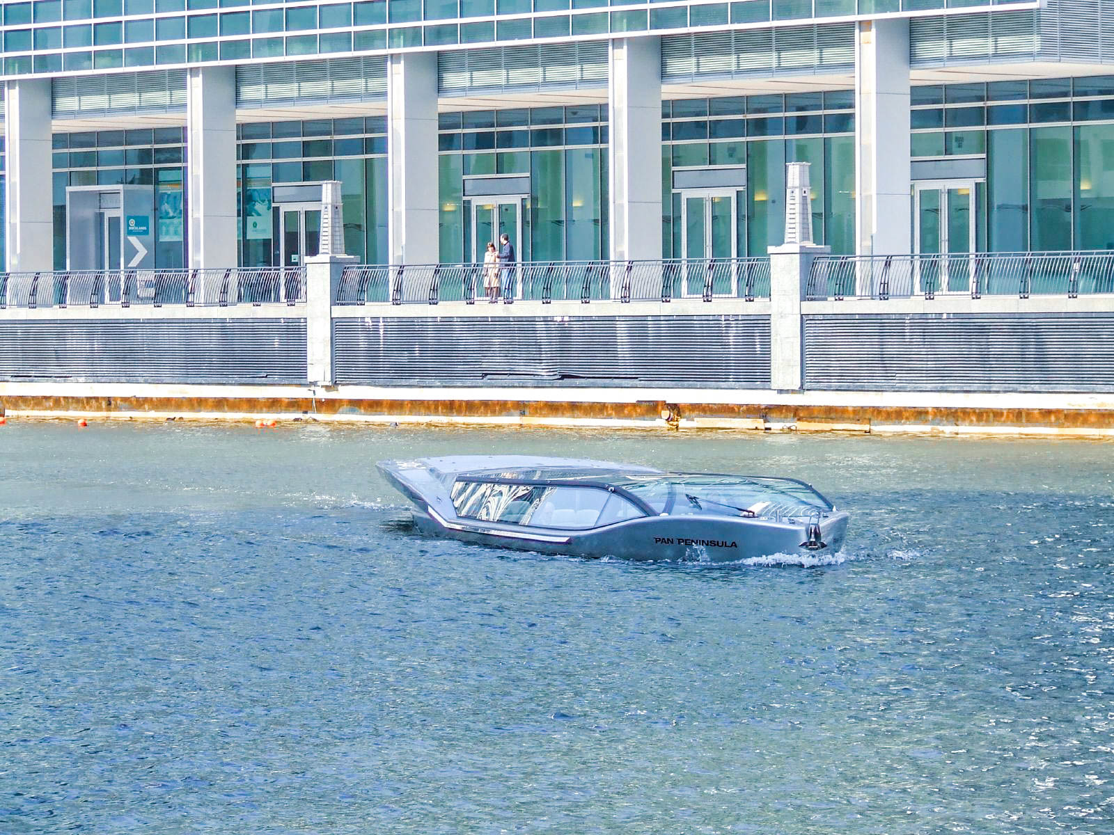 The image shows a sleek, modern amphibious vehicle labeled 'Pan Peninsula' navigating through water in an urban setting. In the background, there is a contemporary building with large glass windows and a few people standing on a walkway near the building.
