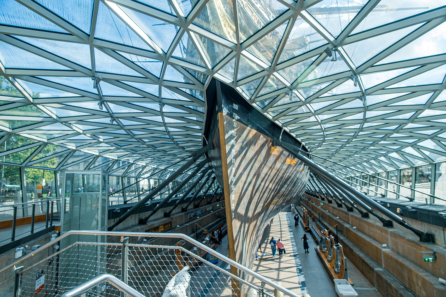 The image depicts a modern, architecturally impressive structure with a glass and steel roof. The interior features a long, straight walkway with railings on both sides, leading to a distant exit. The ceiling is composed of intersecting steel beams and glass panels, allowing natural light to illuminate the space. On either side of the walkway, there are various exhibits, possibly boats or ships, displayed in what appears to be a museum setting. The overall atmosphere is bright and spacious, with a few people walking along the path.