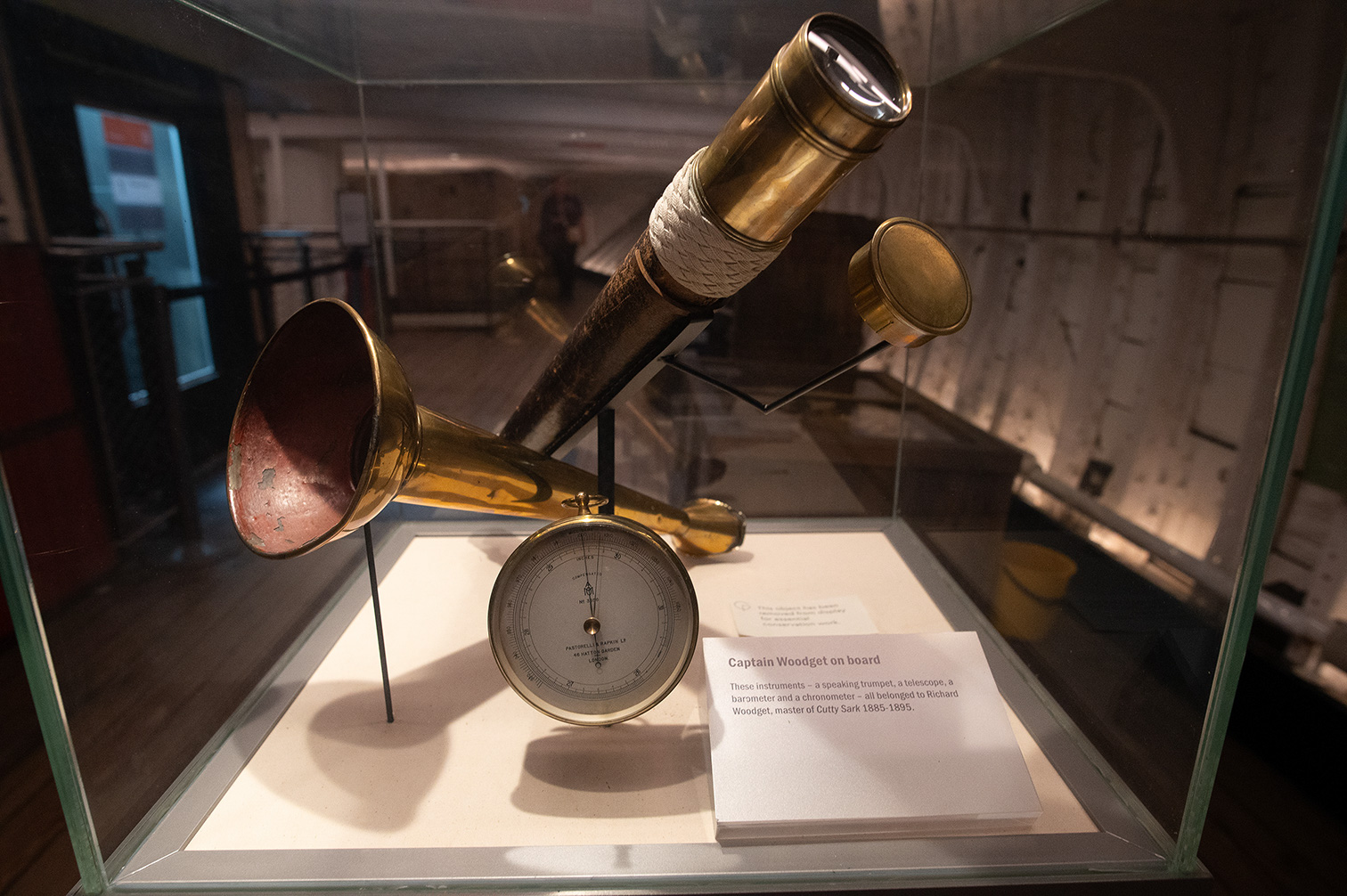 The image shows a display case containing historical navigation instruments, including a speaking trumpet, a telescope, a barometer, and a chronometer. These items belonged to Captain Richard Woodget, who was the master of the ship Cutty Sark from 1885 to 1895.