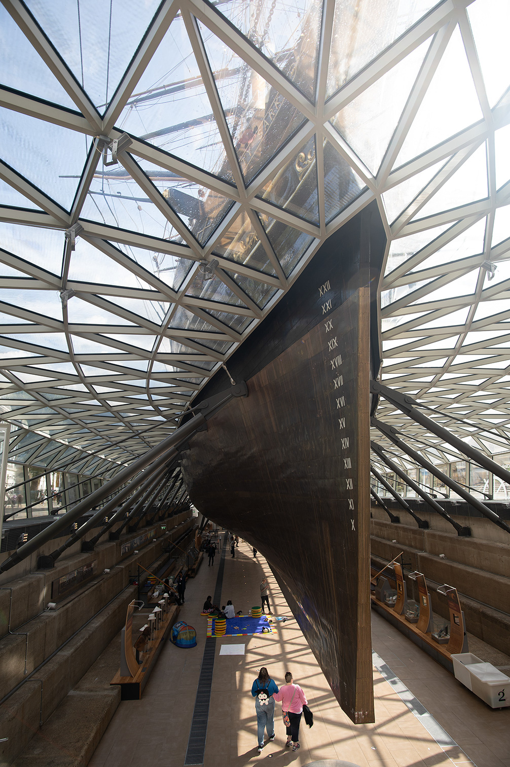 The image depicts an indoor museum exhibit featuring a large, rusted ship's anchor on display. The anchor is mounted vertically and has Roman numerals etched into it, indicating its historical significance. The exhibit is housed in a modern, glass-covered structure with a geometric, lattice-like roof that allows natural light to illuminate the space. Visitors can be seen walking around and observing the anchor and other maritime artifacts displayed in the museum.