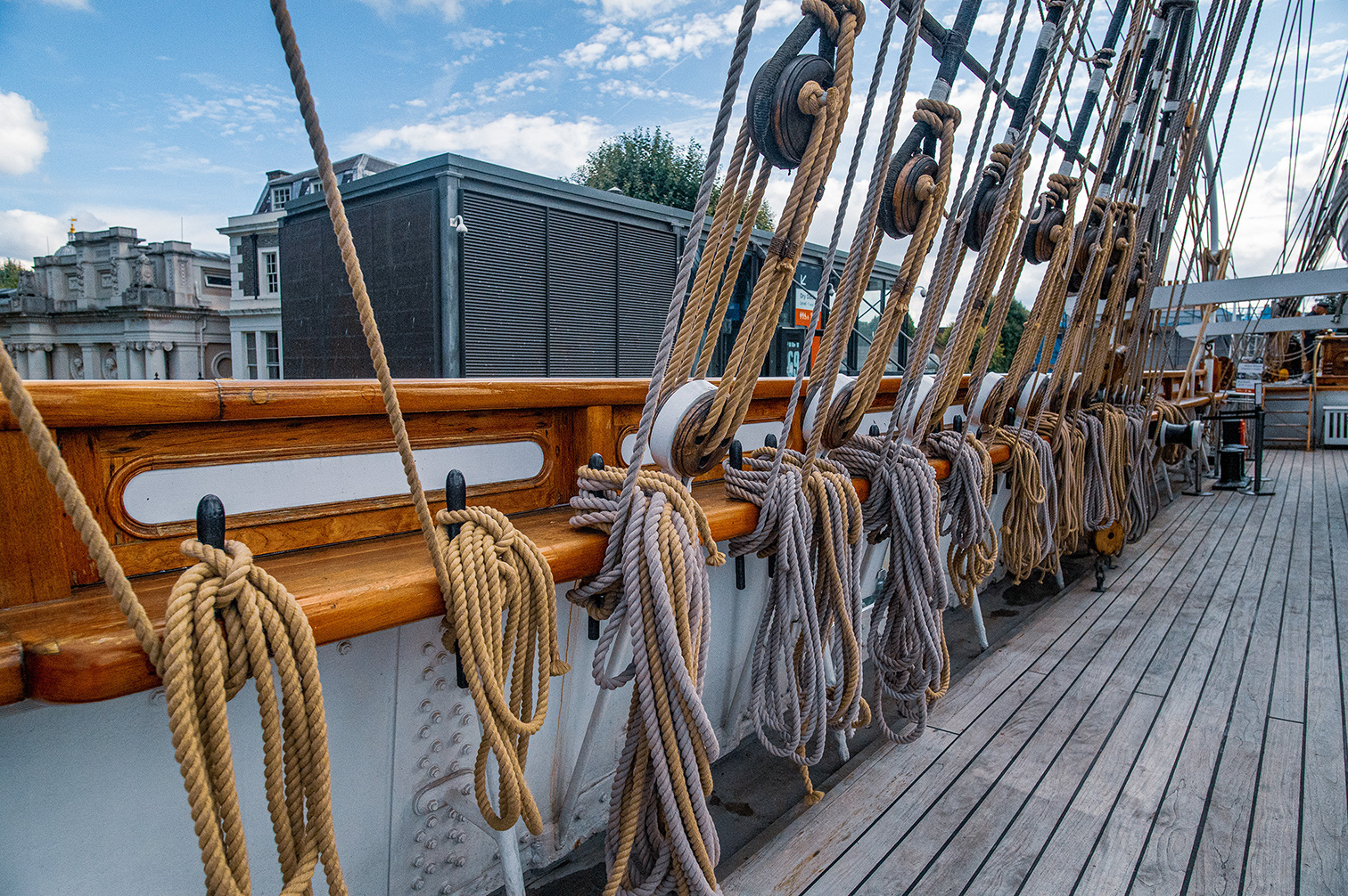 The image depicts the deck of a wooden ship, showcasing various ropes, pulleys, and wooden railings. The ship appears to be docked, with modern buildings visible in the background. The deck is well-maintained, with neatly arranged ropes and pulleys, indicating it might be part of a historical or tourist attraction.