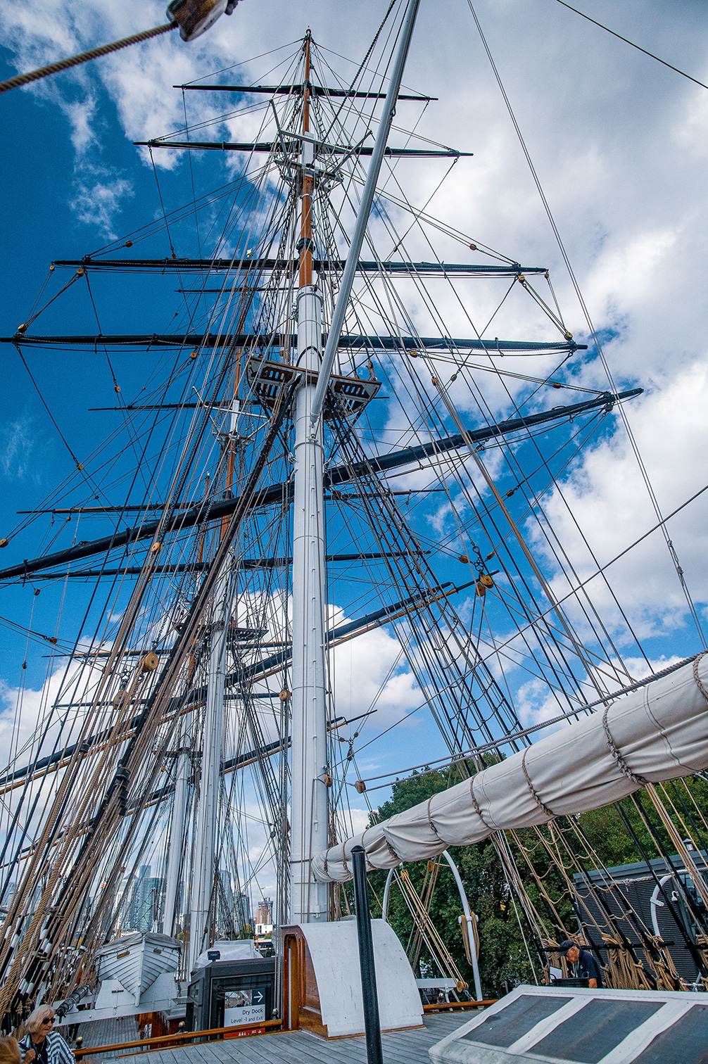 The image depicts the rigging and masts of a large sailing ship against a partly cloudy sky. The perspective is from the deck, looking upwards, showcasing the intricate network of ropes and spars. The ship appears to be docked, with parts of the deck and other structures visible. The overall scene conveys a sense of the ship's impressive size and the complexity of its rigging.