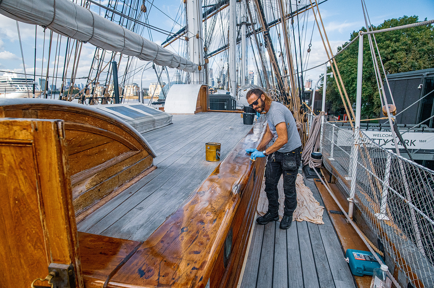 A man is diligently cleaning and polishing the wooden surfaces of a ship's deck. He is wearing a grey t-shirt, jeans, and sunglasses, and is using a cloth and what appears to be a cleaning solution. The ship is equipped with various ropes, rigging, and sails, and is docked near a modern building and a body of water. The scene suggests a blend of historical and contemporary elements.