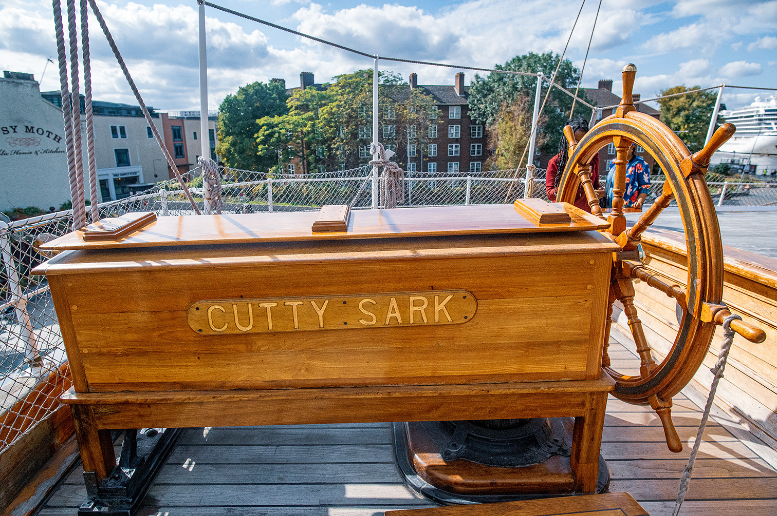 The image shows the stern of a ship named 'Cutty Sark,' featuring its wooden structure, large ship's wheel, and the nameplate prominently displayed. The background includes a view of buildings and trees, suggesting the ship is docked in an urban area with a mix of modern and historic architecture.