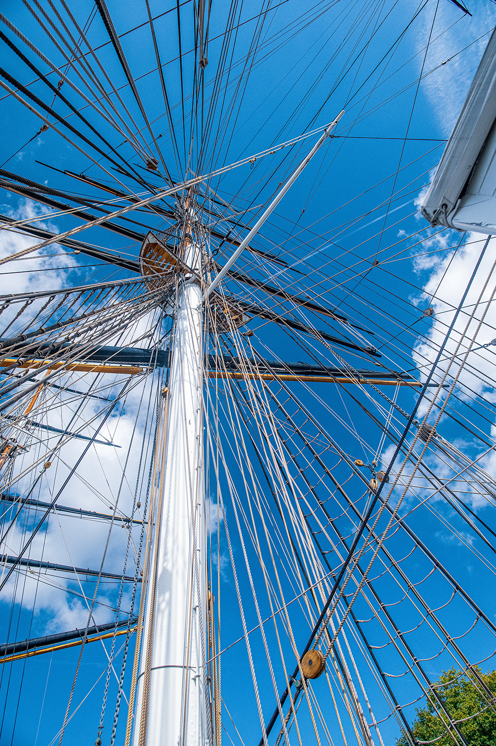 The image depicts the rigging of a tall ship against a blue sky with scattered clouds. The view is from below, looking upwards along the mast, showing a complex network of ropes and lines extending in various directions. The intricate web of lines and the wooden pulleys are clearly visible, highlighting the detailed and elaborate structure of the ship's rigging.