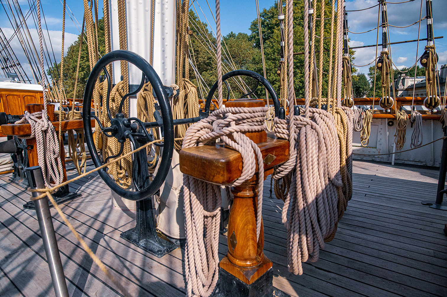 The image depicts the deck of a historical sailing ship. Visible are the ship's wheel, wooden railings, and various ropes and pulleys used for operating the sails. The deck is made of wooden planks, and the background shows masts and rigging against a partly cloudy sky with trees visible in the distance.