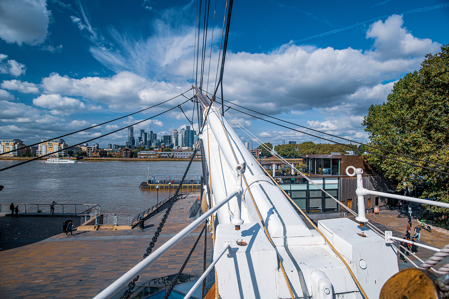 The image depicts a view from the deck of a ship, likely a sailboat, looking out over a river or harbor. The ship's rigging and masts are prominently featured in the foreground. In the background, there is a cityscape with modern buildings and skyscrapers, suggesting an urban setting. The sky is mostly clear with some clouds, and the overall atmosphere appears to be bright and sunny. People can be seen walking along the waterfront, indicating a bustling and lively environment.