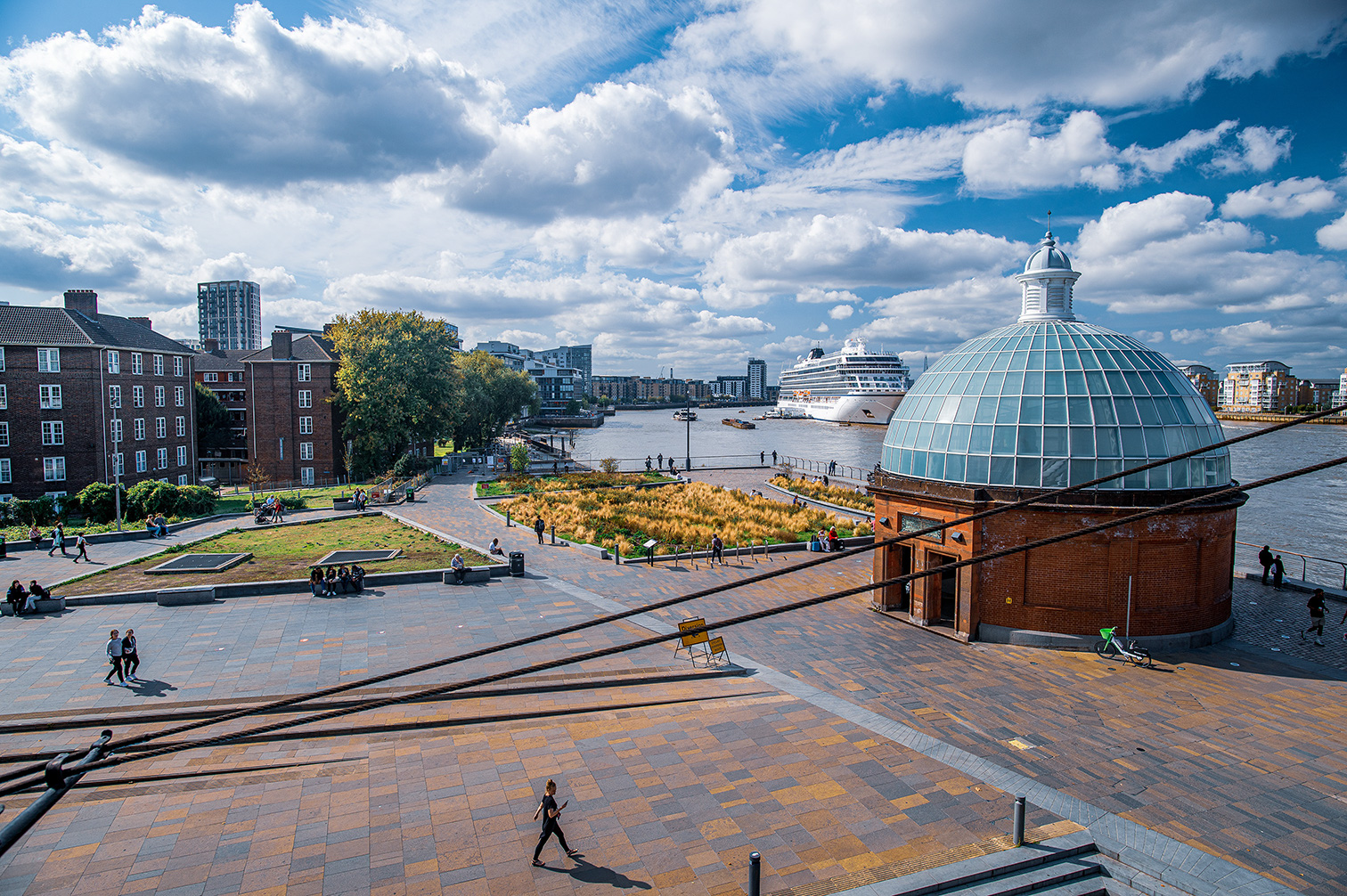 The image depicts a scenic urban waterfront area with a large, dome-shaped structure in the foreground. The area features paved walkways, green spaces, and people leisurely walking and sitting. In the background, a large cruise ship is docked along the river, surrounded by modern buildings and a mix of older brick structures. The sky is mostly clear with some scattered clouds, suggesting a pleasant day.