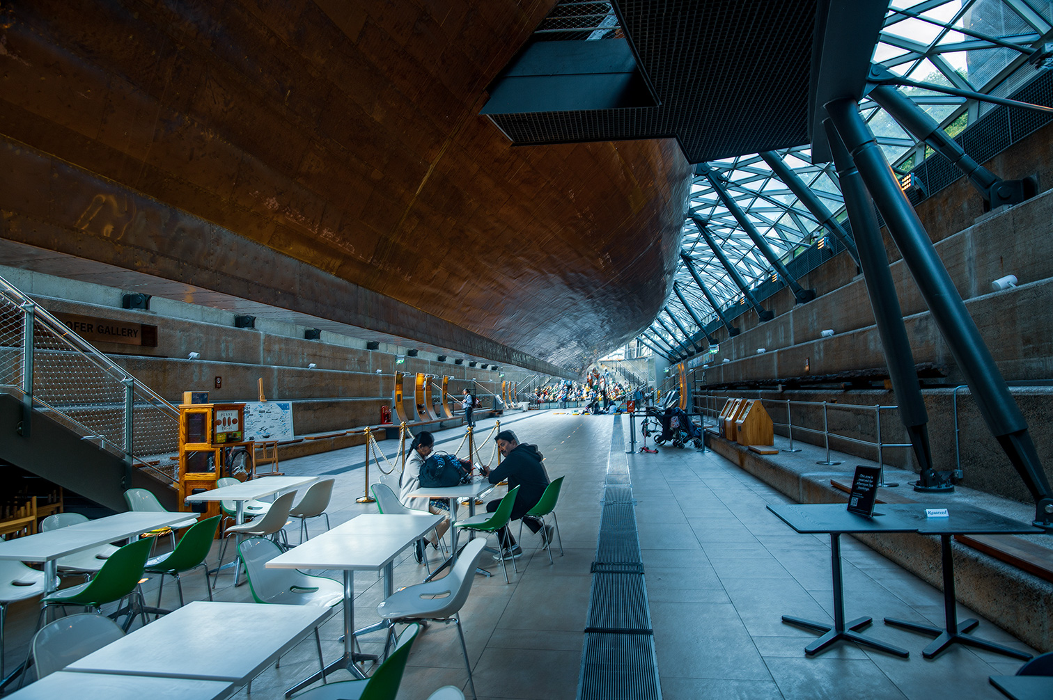 The image depicts an indoor space with a high, arched ceiling made of wood and glass, featuring a series of tables and chairs arranged in a modern, minimalist style. The area appears to be a part of a public building, possibly a gallery or a station, as indicated by the signage and the presence of informational displays. People are seated at the tables, engaged in various activities such as working or studying. The overall atmosphere is bright and spacious, with natural light streaming in through the glass ceiling.