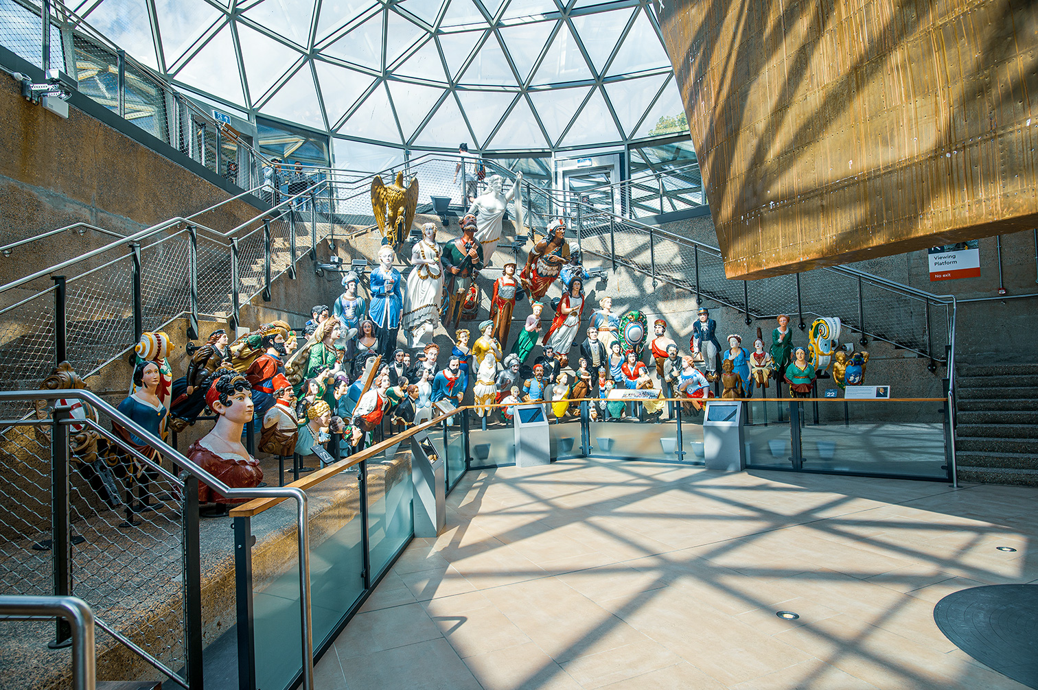The image depicts a staircase with a collection of mannequins dressed in various historical and cultural costumes. The mannequins are arranged in a tiered fashion, creating a visually striking display. The setting appears to be within a modern structure with a glass ceiling, allowing natural light to illuminate the scene. The mannequins are positioned on both the stairs and the landings, adding depth and dimension to the display. The overall atmosphere is one of a museum or exhibition space, showcasing a diverse array of attire and historical representations.