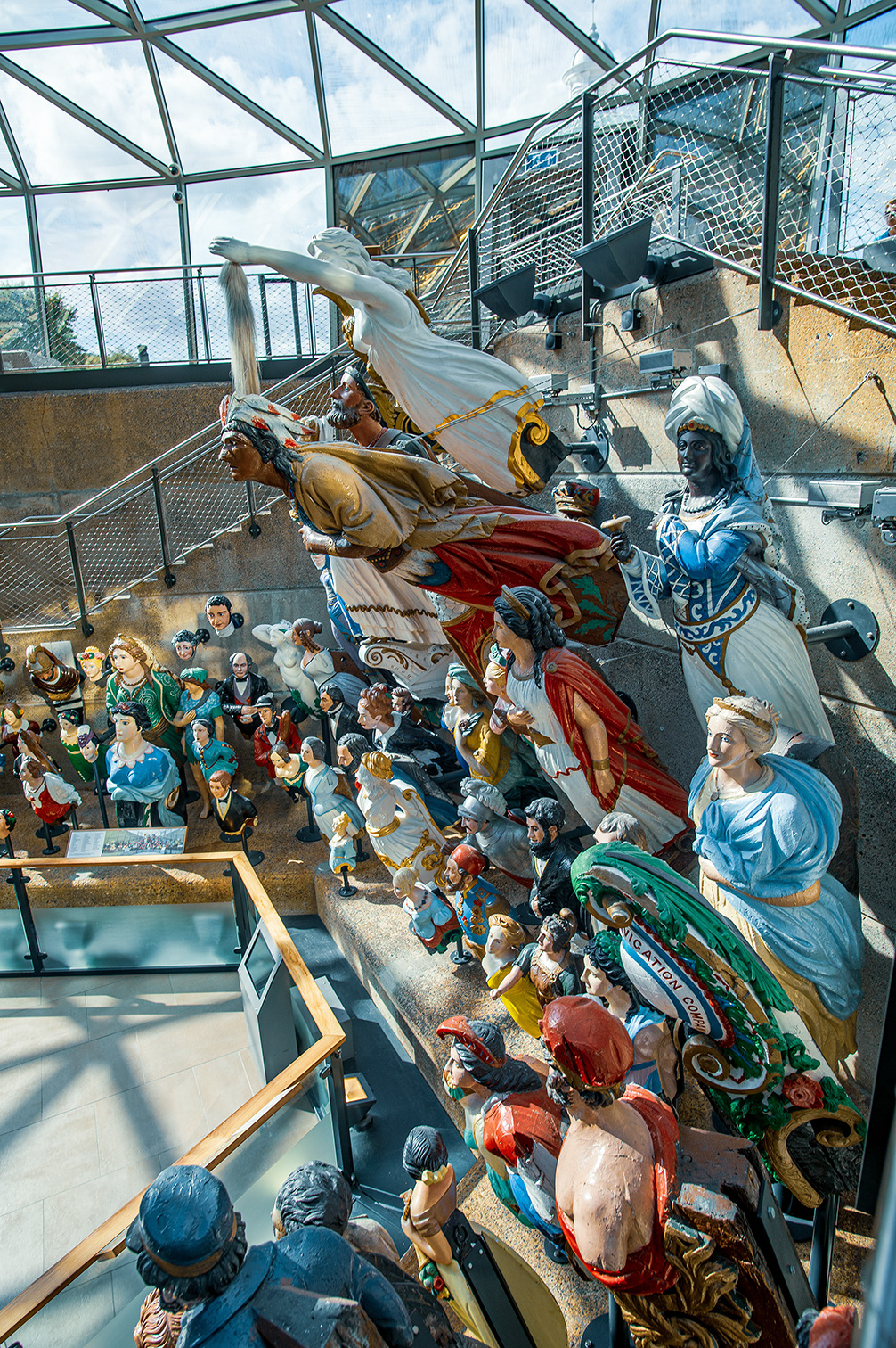The image depicts a detailed and colorful diorama or model scene, likely a historical or biblical representation, featuring numerous figurines in elaborate costumes and settings. The scene is housed in a modern structure with glass and metal elements, suggesting it is part of an indoor exhibit or museum display.