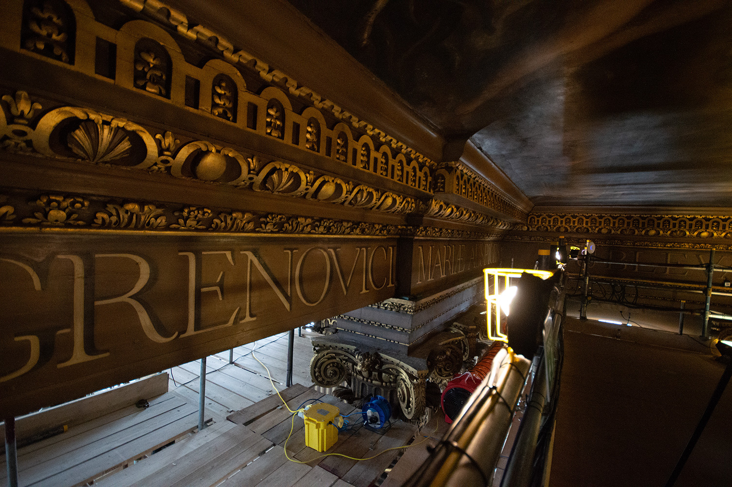 The image shows an ornate interior space, likely a historic or religious building, undergoing restoration or maintenance work. Scaffolding and lighting equipment are set up, with workers possibly present. The visible text 'RENOVICI MARY A' suggests a connection to renovation activities. The intricate carvings and architectural details indicate a richly decorated ceiling.