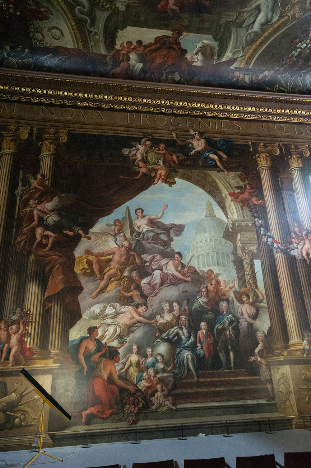 The image depicts a detailed fresco with classical elements, featuring numerous figures in various poses and attire, set against an architectural backdrop. The scene includes a central figure pointing towards a domed structure resembling the U.S. Capitol building. Surrounding figures appear to be in states of reverence or offering, with cherubs and other classical motifs scattered throughout. The fresco is framed by ornate columns and an arch, with a Latin inscription at the top.