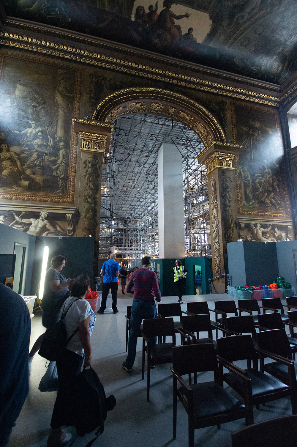 The image depicts an interior of a grand, ornate building under renovation. Scaffolding is visible, and workers are present, indicating ongoing restoration work. The space features large, detailed paintings and classical architectural elements. Visitors are seen observing the work, suggesting that the site is accessible to the public despite the renovation.