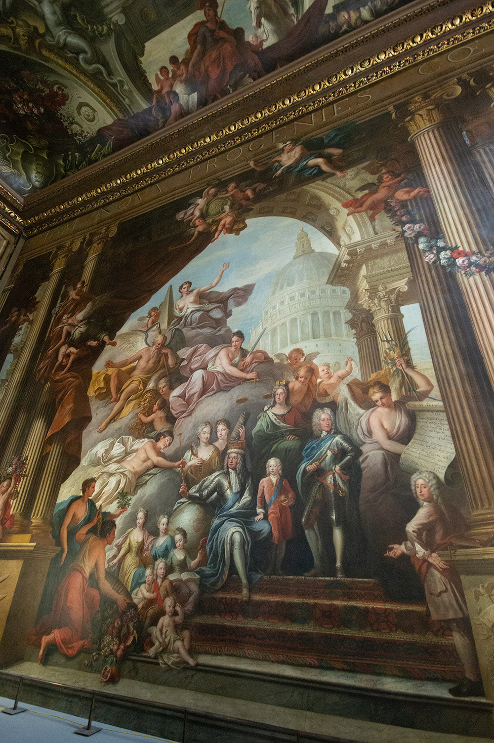 The image depicts a grand, ornate ceiling fresco featuring numerous figures, including angels, mythological characters, and historical individuals. The central figure appears to be a winged entity, possibly representing a deity or allegorical figure, surrounded by a group of people in various poses. The background includes architectural elements like columns and a dome, suggesting a classical or neoclassical style. The fresco is rich in detail, with intricate gold accents and a variety of colors enhancing the overall grandeur.