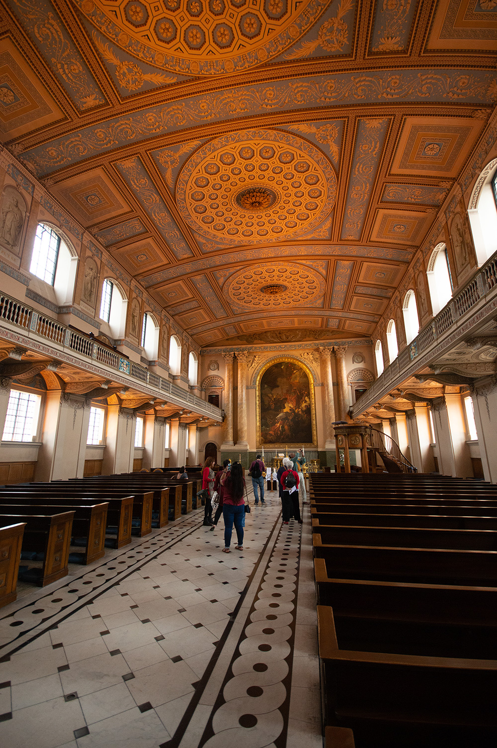 The image depicts the interior of a grand, ornate building, likely a church or cathedral. The ceiling features intricate, geometric patterns and detailed artwork. The space is filled with wooden pews arranged in rows, and there are balconies on either side. A group of people is seen walking down the central aisle, and a large, elaborate painting is visible at the far end of the room.