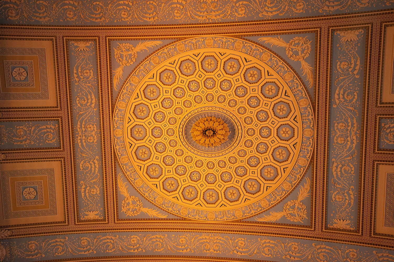 The image showcases an intricately designed ceiling with a central circular pattern composed of geometric shapes and floral motifs. The design is symmetrical and features a radial pattern that extends outward from the center. The surrounding areas include detailed ornamental patterns and classical motifs, such as Greek keys and floral decorations, all rendered in a rich, golden color palette.