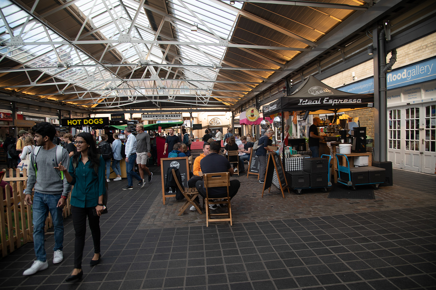 The image depicts a bustling indoor market with various food stalls, including a hot dog stand and an espresso bar. The market has a high, glass-paneled ceiling allowing natural light to illuminate the space. People are seen walking around, sitting, and enjoying their meals. The market appears to be a lively and popular spot for socializing and dining.