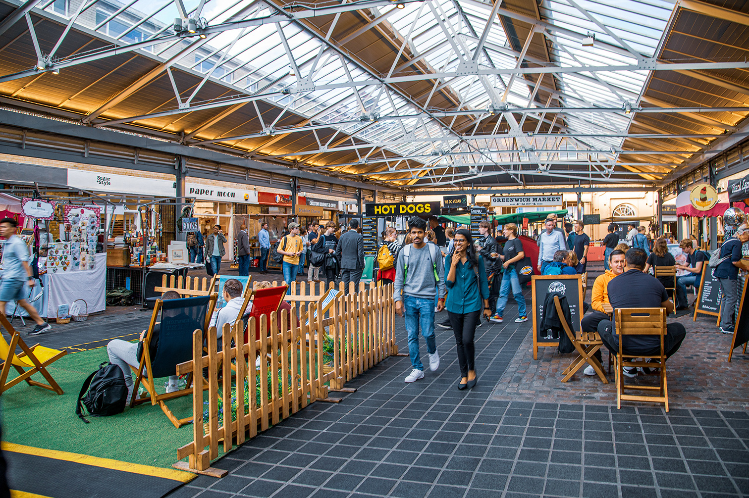 The image depicts a bustling indoor market with various stalls and shops. The market features a high, glass-paneled roof allowing natural light to illuminate the space. People are seen walking, shopping, and conversing, creating a lively atmosphere. There are seating areas with wooden chairs and tables where individuals are sitting and relaxing. The stalls offer a variety of goods, including books, clothing, and food items like hot dogs.