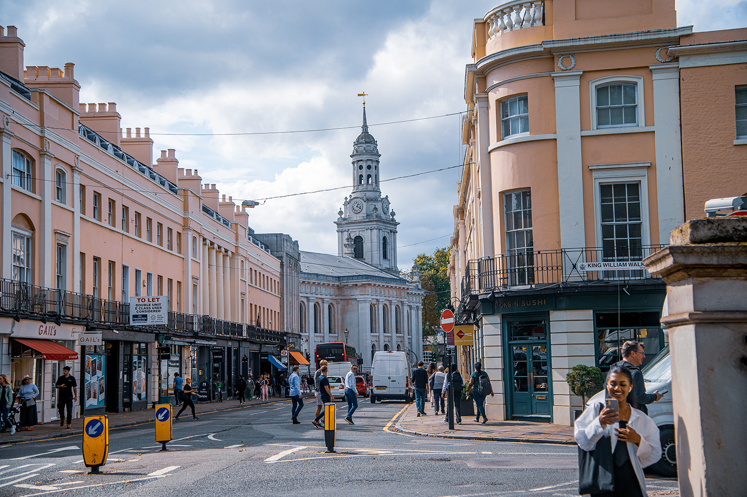 The image depicts a bustling street scene in an urban area with a mix of pedestrians and vehicles. The street is lined with pastel-colored buildings, shops, and cafes. A prominent church with a tall clock tower is visible in the background. The sky is partly cloudy, and the overall atmosphere appears lively and busy.