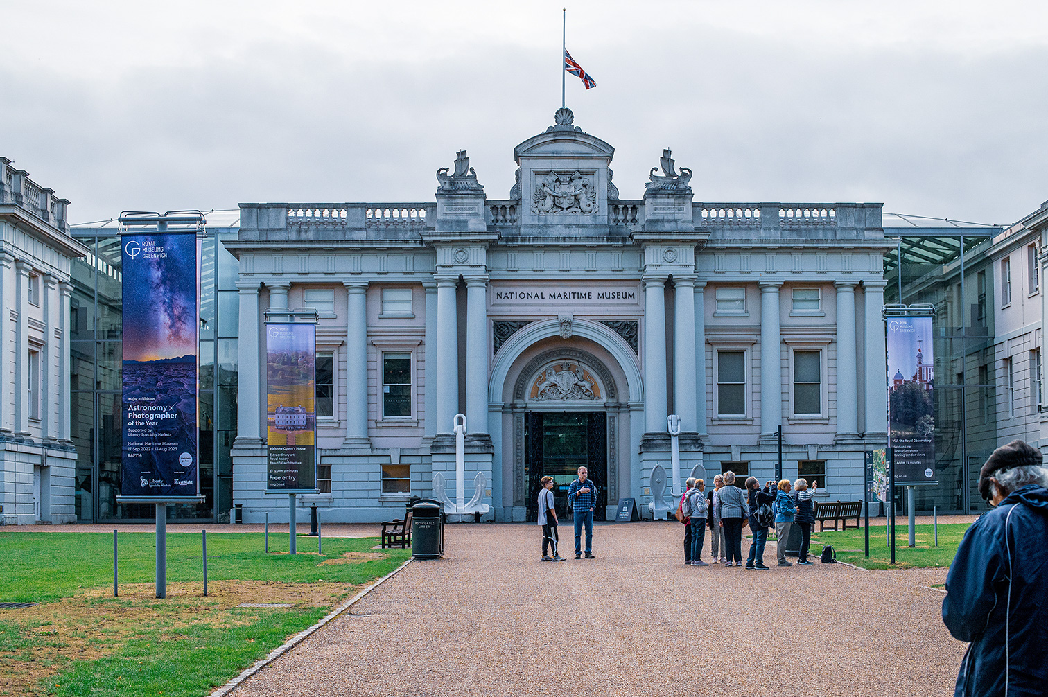 Visitors gather at the entrance of the National Maritime Museum, a stately building featuring prominent columns and classical architecture. Banners promoting exhibitions, including &lsquo;Astronomy x Photographer of the Year&rdquo; and &lsquo;Visit the Queen's House,&rdquo; line the pathway. Large anchor sculptures flank the main entrance, adding to the maritime theme. The scene is set against a cloudy sky, with a Union Jack flag flying from the rooftop.