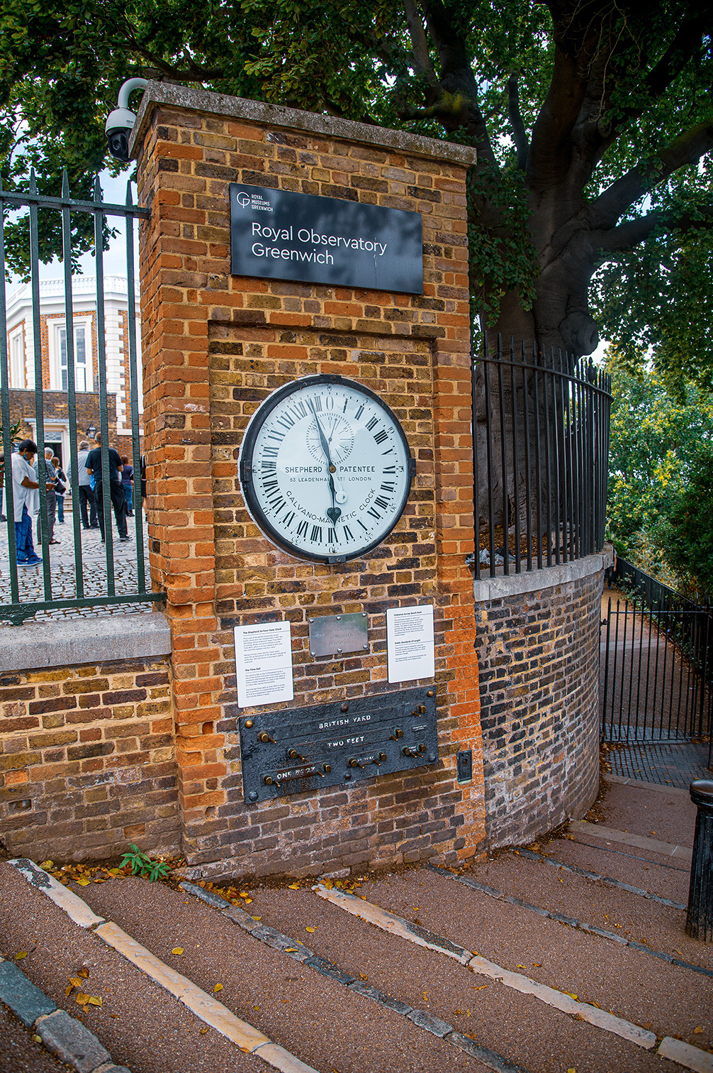 The image shows the entrance to the Royal Observatory in Greenwich, London. The entrance is marked by a brick pillar with a sign reading 'Royal Observatory Greenwich' and a clock displaying the time. Below the clock, there is a plaque indicating the 'British Yard' and 'Two Feet' measurements. The area is surrounded by a black iron fence, and there are informational placards on the pillar. The scene is set in a green, leafy environment with a few people visible in the background.