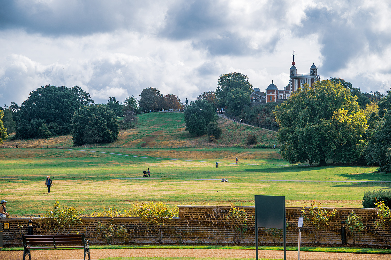 The image depicts a large, open park with a few people walking and relaxing. The park features a gently sloping grassy area with scattered trees. In the background, there is a large, historic-looking building with a prominent tower and multiple domed structures. The sky is overcast, and there is a brick wall with benches and a sign in the foreground.