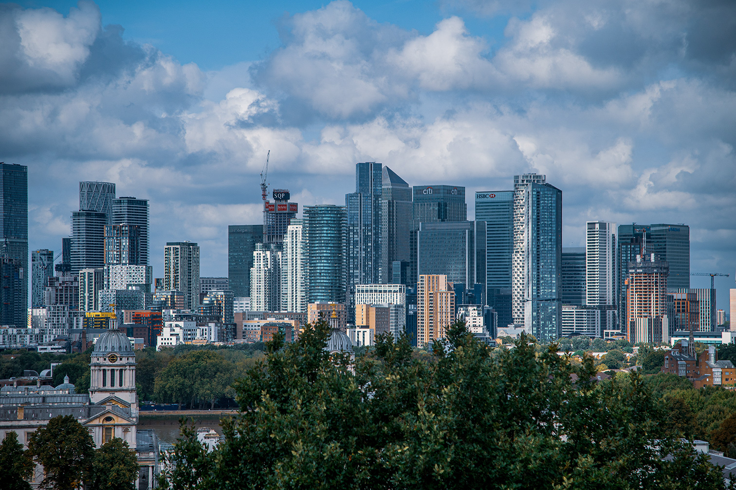 The image depicts a cityscape featuring a mix of modern skyscrapers and older, classical buildings. The skyline is dominated by tall, glass-clad office buildings with recognizable logos of major banks such as Citi and HSBC. In the foreground, there are lush green trees and classical architecture, providing a contrast to the contemporary high-rises in the background. The sky is partly cloudy, adding a dynamic element to the scene.