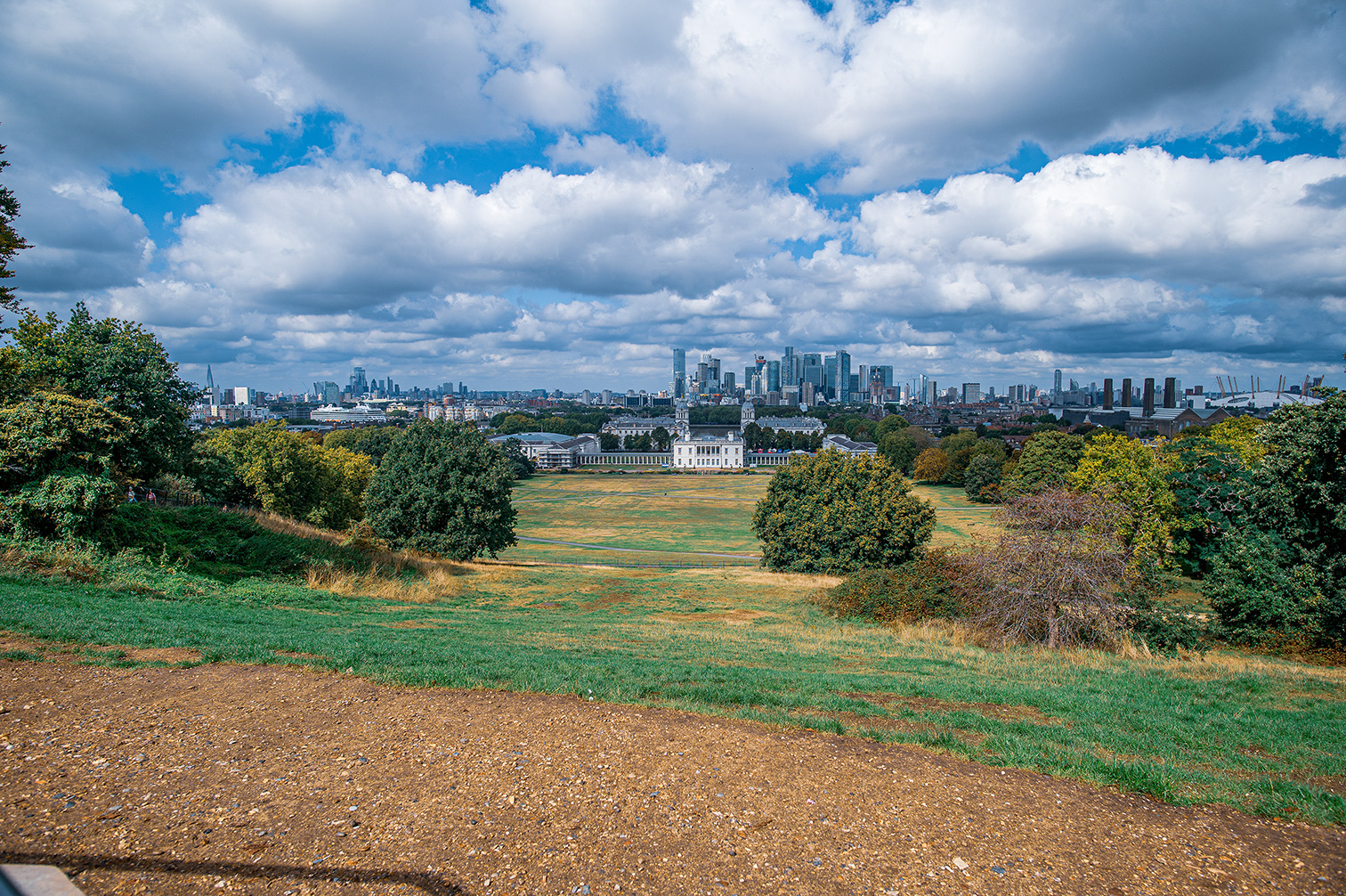 The image depicts a scenic view of a park with lush greenery and a clear path leading towards a historic building in the distance. Beyond the building, the skyline of a modern city with tall skyscrapers is visible under a cloudy sky.