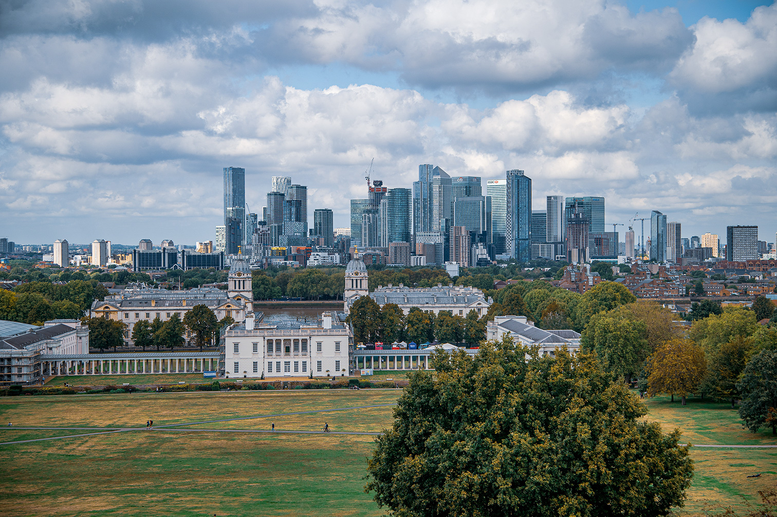 The image depicts a cityscape with a mix of modern skyscrapers and historical buildings. In the foreground, there is a large, open green space with a few people walking. The middle ground features historical buildings with classical architecture, including domed structures. The background showcases a cluster of modern skyscrapers, indicating a bustling urban area.