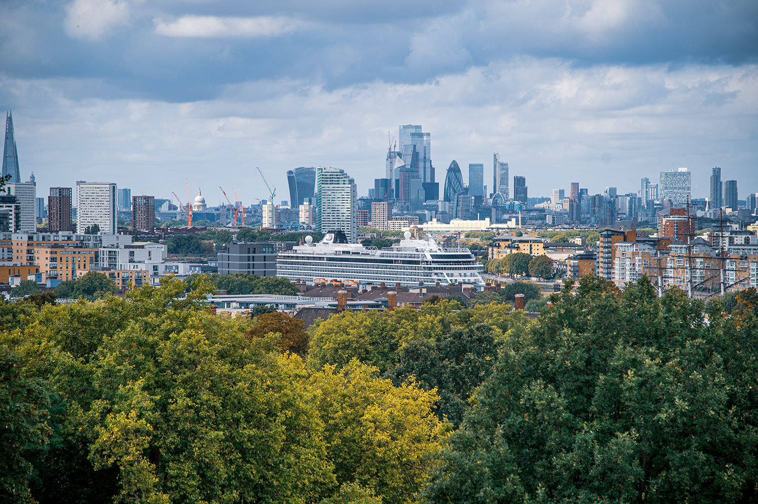The image depicts a cityscape with a mix of modern skyscrapers and older buildings, set against a backdrop of a cloudy sky. In the foreground, there is a lush expanse of greenery, including trees with green and yellow foliage, suggesting a park or natural area. The city skyline features notable architectural structures, including the distinctive Gherkin and Walkie Talkie buildings, as well as the Shard. There are several construction cranes visible, indicating ongoing development. The overall scene combines urban density with natural elements, showcasing a vibrant and dynamic city environment.