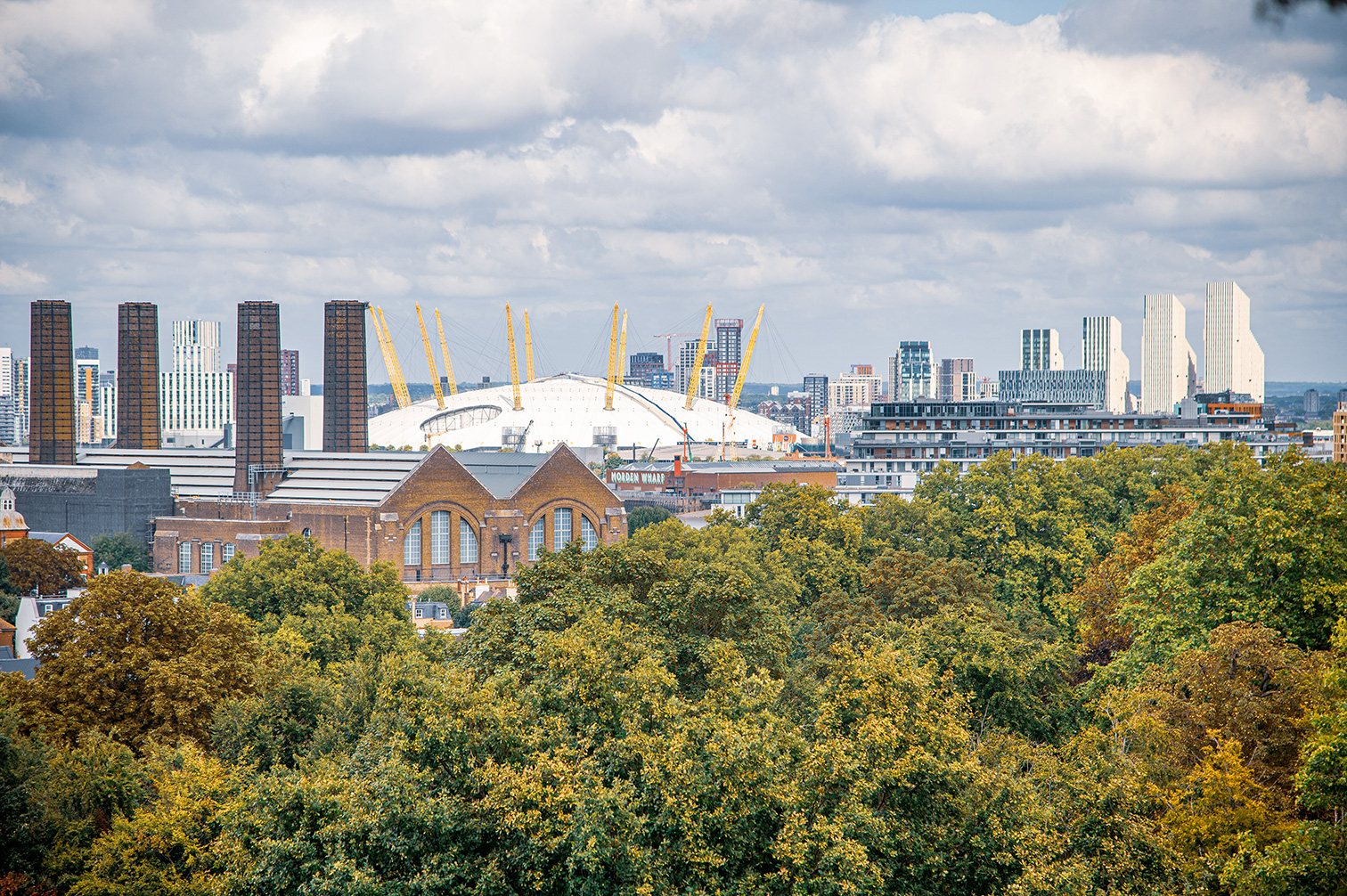 The image depicts a cityscape featuring a mix of modern and historic architecture. In the foreground, there is a lush expanse of green trees. Prominently visible in the middle of the image is a large, white dome-shaped structure with yellow support beams, identified as the O2 Arena. Surrounding this structure are various buildings, including tall, modern skyscrapers and older, brick buildings. The sky above is partly cloudy, adding to the overall urban atmosphere of the scene.