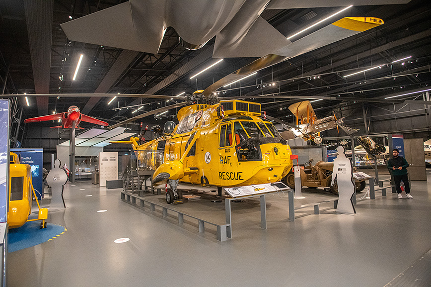 The image depicts an aviation museum exhibit featuring a prominent yellow Royal Air Force (RAF) rescue helicopter on display. Surrounding the helicopter are various other aircraft and aviation-related exhibits, including a red plane suspended from the ceiling and a small yellow vehicle. The museum setting includes informational displays and interactive elements, with visitors observing the exhibits.
