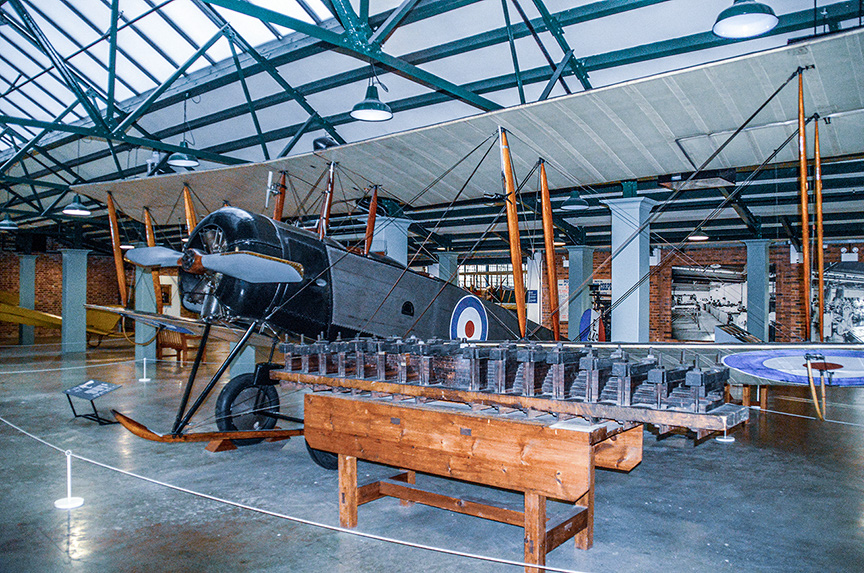 The image depicts an indoor aviation museum exhibit featuring a vintage military aircraft with a roundel insignia on its side. The aircraft is displayed alongside various engine parts and components, likely from the same era, arranged on wooden tables. The museum has a high ceiling with a glass roof, allowing natural light to illuminate the exhibit. The setting suggests a historical or educational display focused on early aviation technology and machinery.