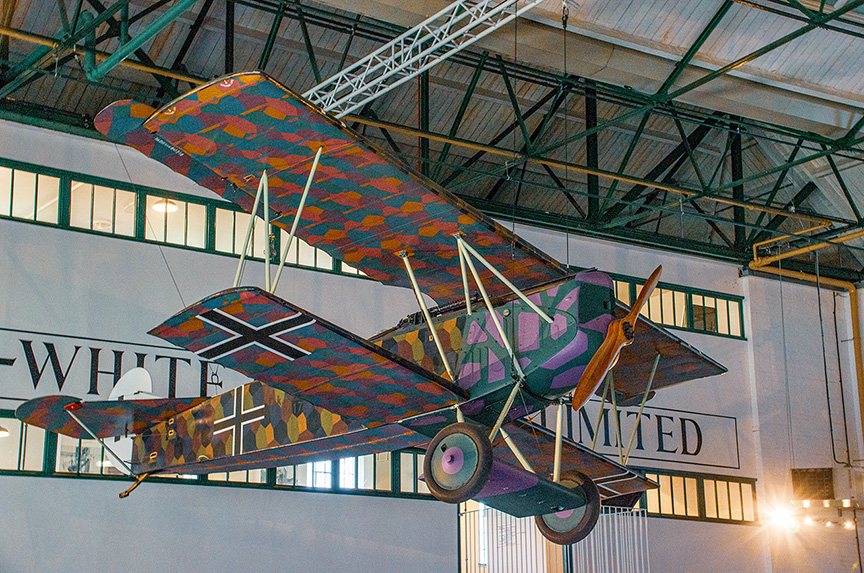 The image depicts a vintage biplane with a colorful, camouflage-like pattern on its wings and body, suspended from the ceiling of a large hangar. The biplane is displayed in front of a sign that reads 'WHITE LIMITED.' The setting appears to be an aviation museum or a similar exhibition space.