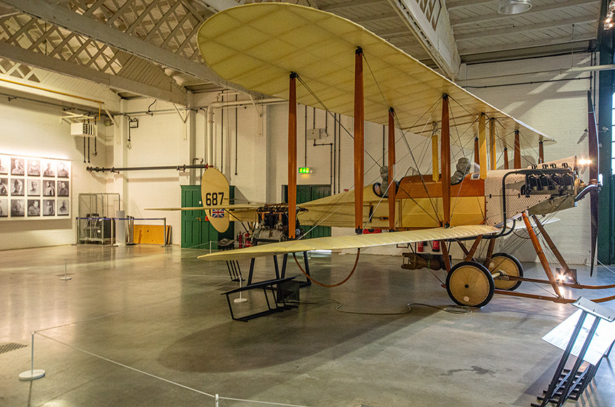 The image depicts an old biplane displayed in a museum setting. The aircraft has a wooden frame and fabric covering, typical of early 20th-century designs. It features a large, prominent tail and two sets of wings, one above the other. The plane is marked with the number 687 and a British flag insignia on the side. The museum space is spacious, with high ceilings and various other exhibits visible in the background.