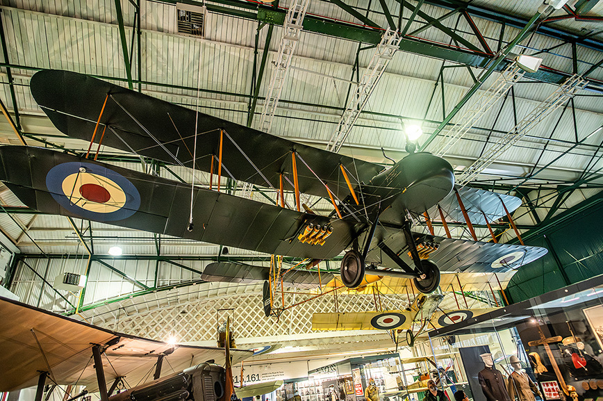 The image shows a vintage military aircraft suspended from the ceiling in a museum setting. The aircraft features a dark green body with yellow markings on the side and circular roundels on the wings. The museum appears to house other aircraft and aviation-related exhibits, with a high ceiling and various display areas visible in the background.