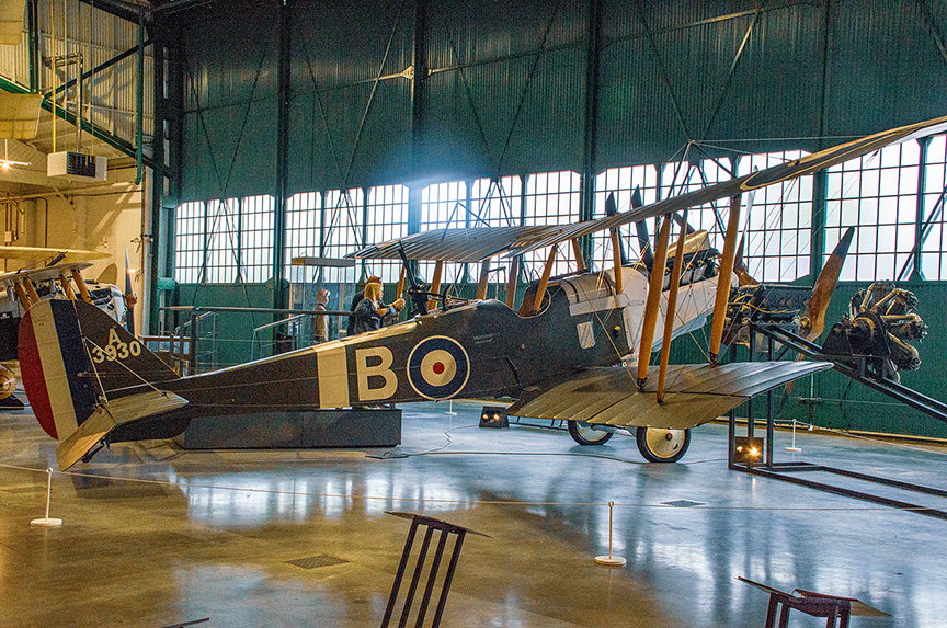 The image shows a vintage biplane displayed in a large hangar or museum setting. The aircraft has a distinctive roundel marking on its side, indicating it is likely of British origin. The plane is mounted on a stand, with its wings spread out, showcasing its biplane design with two sets of wings. The environment is well-lit with natural light coming through large windows, and there are other aircraft visible in the background. The setting appears to be an aviation museum or a similar exhibition space.