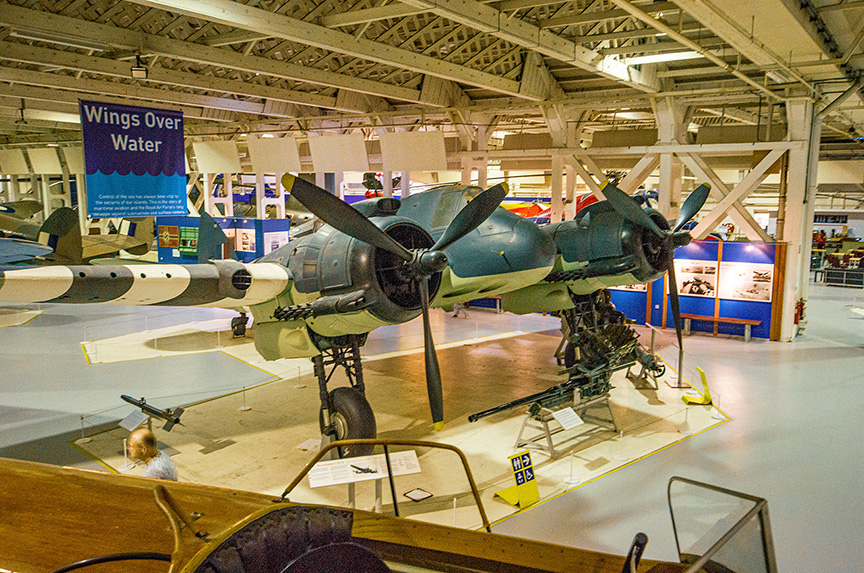 The image shows an aircraft exhibit in a museum. The central focus is on a vintage military aircraft with a distinctive green color scheme and large propellers. The aircraft is displayed on the museum floor with various informational placards around it. In the background, other aircraft and exhibits can be seen, and there is a sign that reads 'Wings Over Water'. The museum setting is spacious with high ceilings and ample lighting.