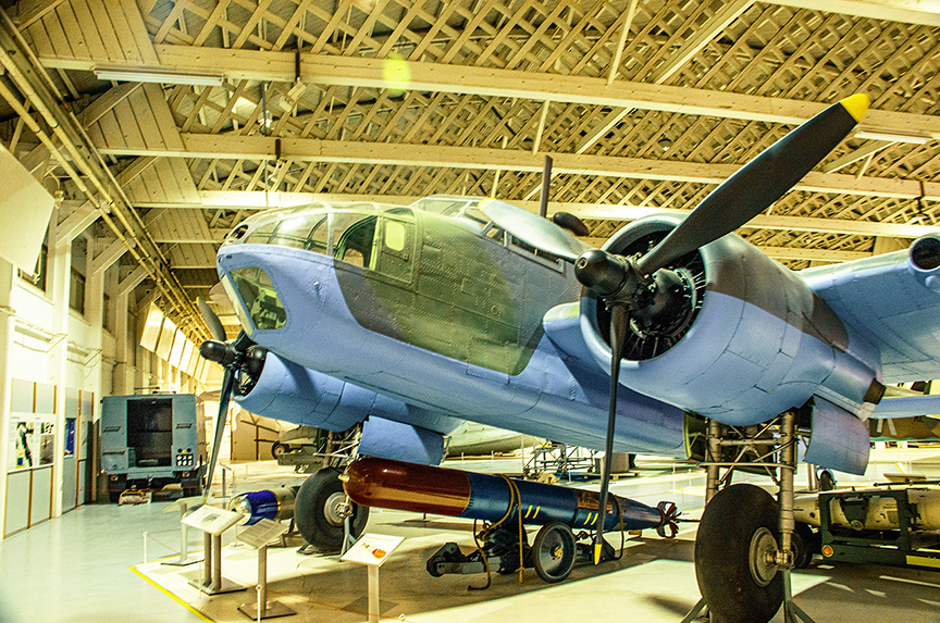 The image shows a vintage military aircraft displayed in a museum. The aircraft is painted in a blue color scheme and features two large propellers. It is mounted on a stand, with informational placards nearby. The setting appears to be an indoor hangar with a high ceiling and wooden beams. The aircraft is equipped with a torpedo or similar weapon mounted underneath.