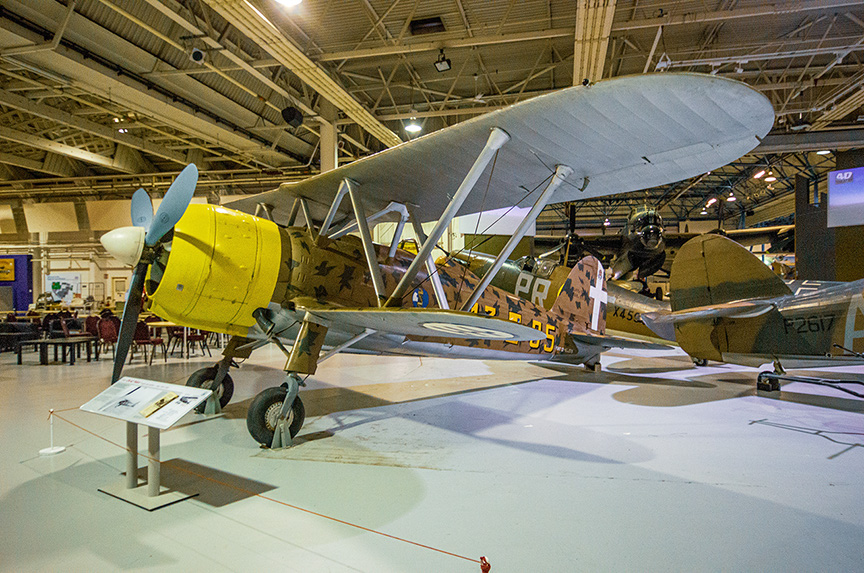 The image depicts a vintage biplane on display in a museum. The aircraft has a yellow propeller and is painted with camouflage patterns on its body. It is positioned on the museum floor with an informational placard in front of it. The museum setting includes other aircraft and exhibits in the background, with visitors seated at tables in the distance.