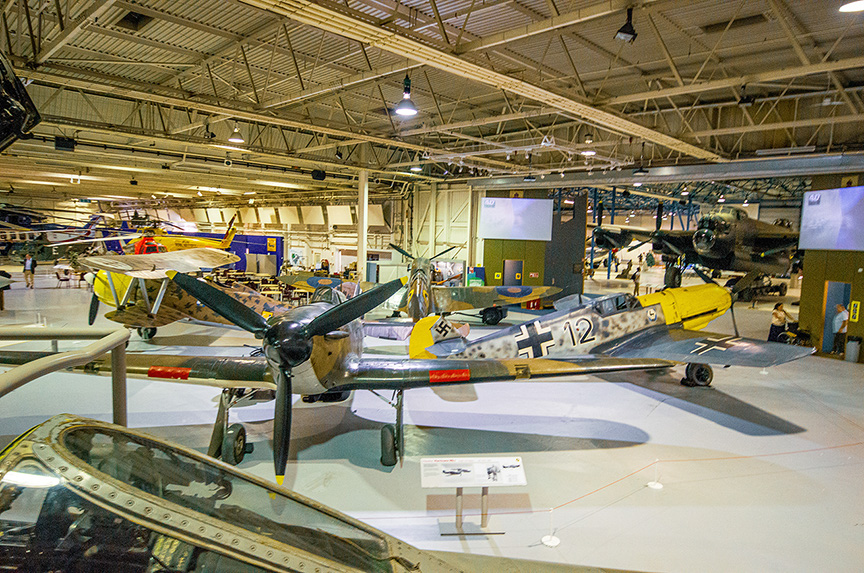 The image depicts an indoor aviation museum with various vintage aircraft on display. The aircraft are arranged in a spacious, well-lit hangar with high ceilings and overhead lighting. The planes are positioned on the floor, with some suspended from the ceiling. Informational placards are placed in front of the aircraft, providing details about each exhibit. The museum appears to be well-organized, with clear pathways for visitors to walk around and view the exhibits.