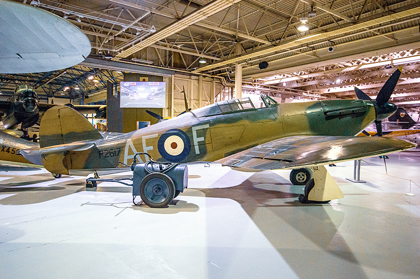 The image shows a vintage military aircraft displayed in a museum setting. The aircraft, likely a World War II fighter plane, is painted in a camouflage pattern with distinctive roundels on its side. The plane is positioned on a stand with its landing gear visible. The museum interior features high ceilings with exposed beams and other aircraft in the background.