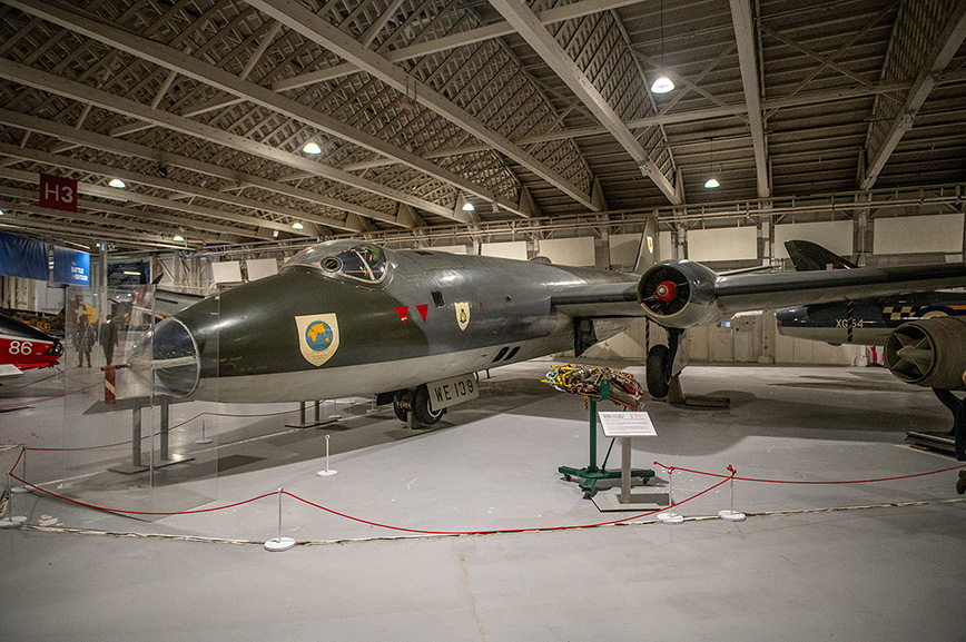 he image shows a military aircraft displayed in a museum. The aircraft is a jet with a sleek, dark green body and distinctive markings on its side. It is positioned indoors under a large, open-roofed structure with high ceilings and industrial lighting. The plane is cordoned off with red ropes and stanchions, indicating it is a protected exhibit. There is a small placard in front of the aircraft, likely providing information about its history and significance.