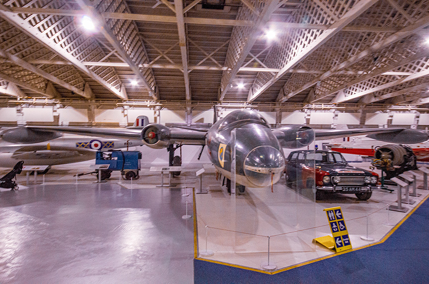 The image depicts an aircraft hangar or museum showcasing various vintage military aircraft. The central focus is on a large, sleek bomber plane with a distinctive nose design, marked with the identification 'C' and 'HX341'. Surrounding this aircraft are other vintage planes and vehicles, including a smaller aircraft with a propeller and a classic car. The hangar has a high ceiling with exposed wooden beams and is well-lit, indicating a preserved and maintained environment for the display of these historical artifacts.