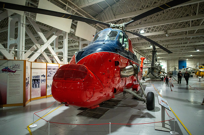The image depicts a red helicopter on display in a museum-like setting. The helicopter is cordoned off with red ropes and stanchions, indicating it is part of an exhibit. The environment is spacious with a high ceiling and industrial lighting, suggesting it is housed in a large hangar or exhibition hall. There are informational displays and other aircraft visible in the background, further emphasizing the museum context. The helicopter appears to be well-maintained and polished, showcasing its vibrant red color and sleek design.