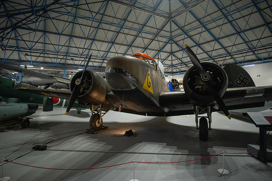 The image depicts a vintage military aircraft, specifically a twin-engine bomber, housed in a large hangar with a distinctive blue truss roof. The aircraft is painted in a dark color scheme with visible markings, including a yellow '6' on the nose and a swastika on the side, indicating it is likely a World War II-era German aircraft. The plane is displayed on a concrete floor with protective barriers around it, suggesting it is part of an aviation museum exhibit.