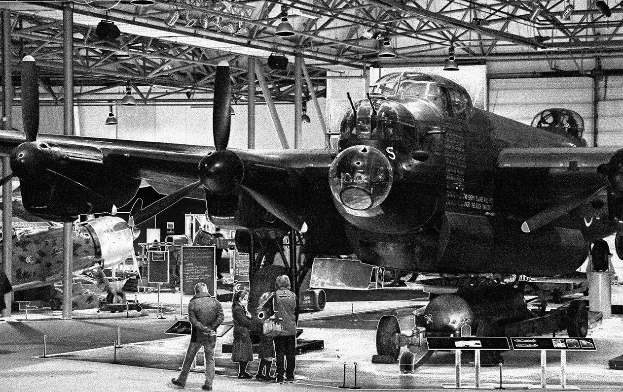 The image depicts an aviation museum exhibit featuring a large, historic bomber aircraft. The aircraft is displayed indoors, with several people observing and discussing it. The museum setting includes other aircraft and informational displays, suggesting an educational environment focused on aviation history.