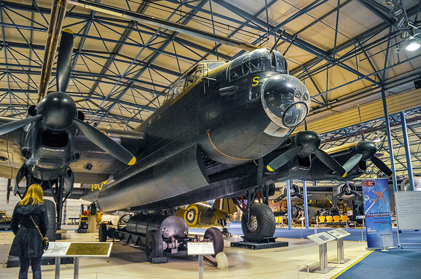 The image shows a large, vintage military aircraft on display in a museum. The aircraft has four large propellers and is painted in a dark color scheme with some yellow markings. The setting appears to be an indoor hangar with high ceilings and other aircraft visible in the background. There are informational placards around the aircraft, and a person is seen observing the display.
